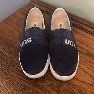 The slip-on suede sneaker features a band of metal charm spelling U-G-G